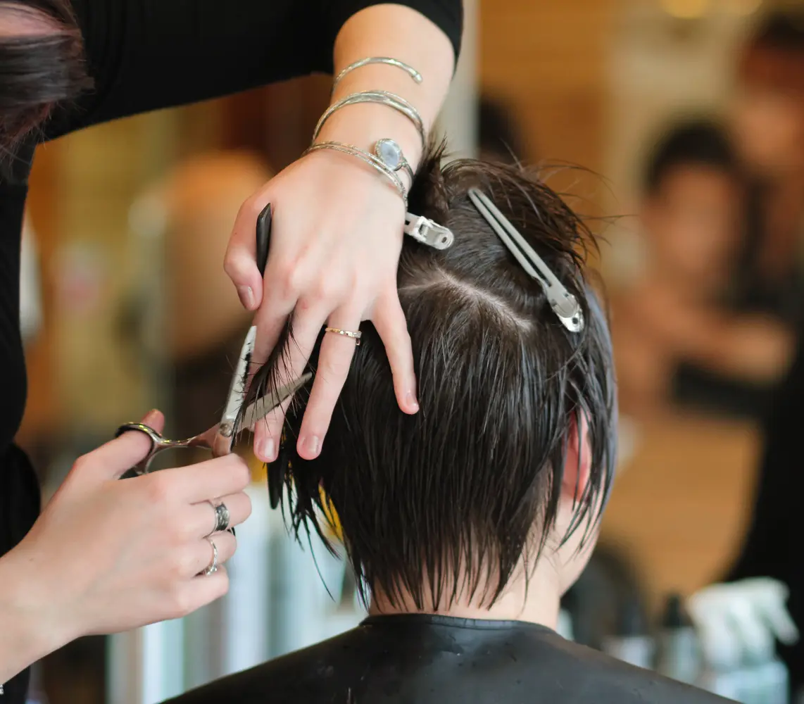 Eine Person mit braunen Haaren wird konzentriert von einem Friseur mit Scheren geschnitten